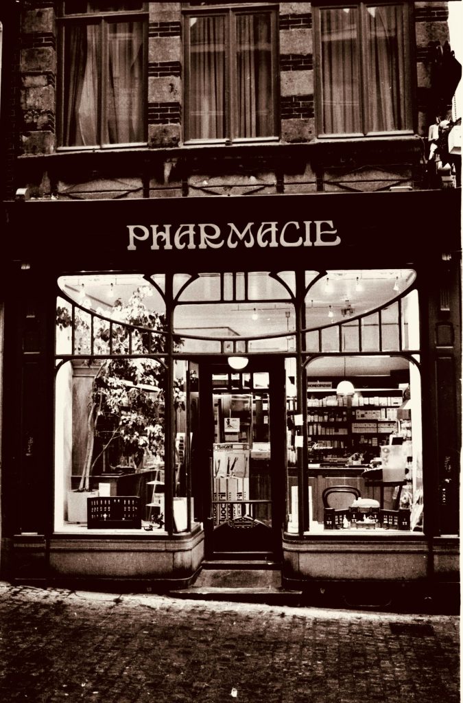 A sepia-toned image of a traditional pharmacy exterior at dusk in Mons, Belgium.